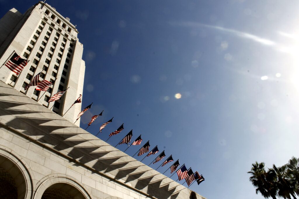 Los Angeles City Hall building rises into a blue sky, with sun shining from the right. American flags hang off the edge of city hall.