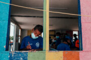 Boy with volunteer shirt writes while standing in a window cut from plywood, painted rainbow colors