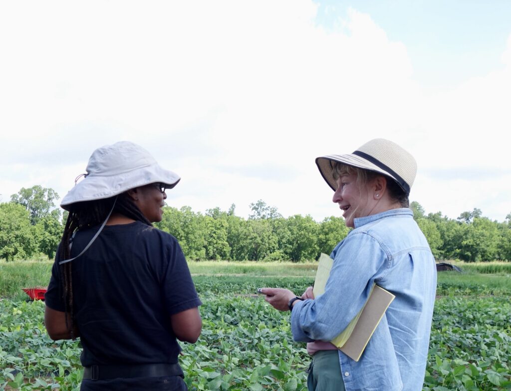 Two women in wide brim hats look at each other while standing in front a field of green with trees in the background. The woman on the left, Konda Mason, is tall and thin, wearing a black t-shirt, and the woman on the right, Diane Winston, carries a pad of paper and recorder in her hand.