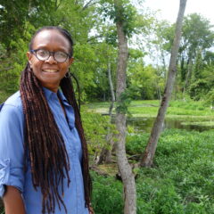 Black woman in glasses and long dreads stands in front of a green trees and a pond.