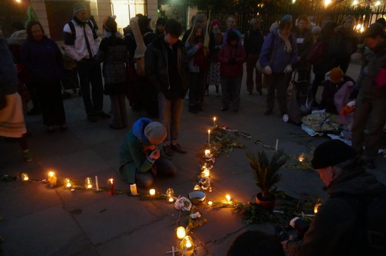 A cross is formed by candles on the ground, one person is kneeling next to the crux of the cross while a large group of people stands around the cross.