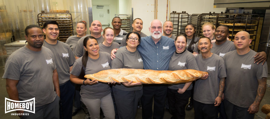 Father Greg and Homeboy Industry trainees smiling and holding freshly baked bread at Homeboy Bakery.